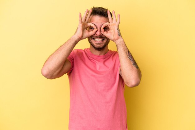 Jovem homem caucasiano isolado no fundo amarelo animado mantendo o gesto okey no olho.