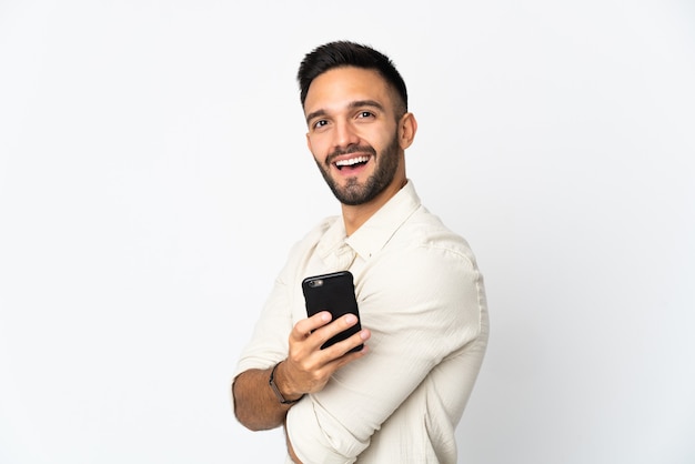 Jovem homem caucasiano isolado no fundo branco segurando um telefone celular e com os braços cruzados