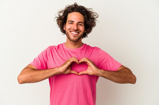 Jovem homem caucasiano isolado no fundo branco, sorrindo e mostrando uma forma de coração com as mãos.