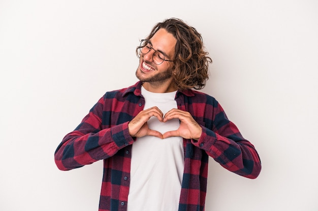 Jovem homem caucasiano isolado no fundo branco, sorrindo e mostrando uma forma de coração com as mãos.