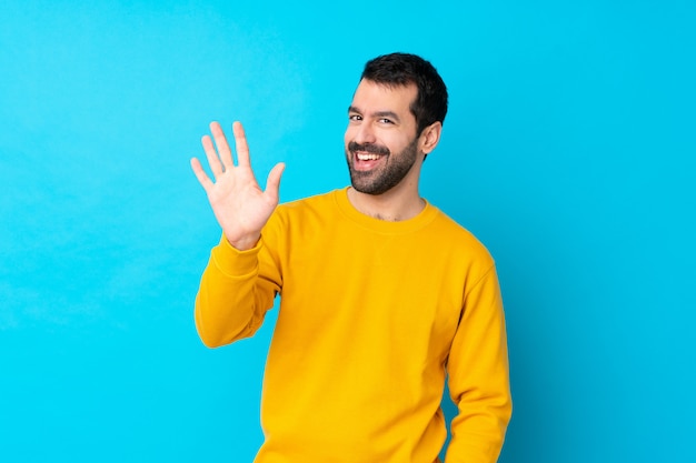 Jovem homem caucasiano isolado parede azul saudando com mão com expressão feliz