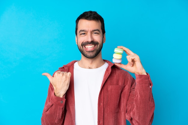 Jovem homem caucasiano isolado parede azul segurando macarons franceses coloridos e apontando o lado