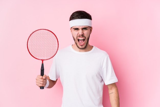 Jovem homem caucasiano jogando badminton isolado gritando muito zangado e agressivo.