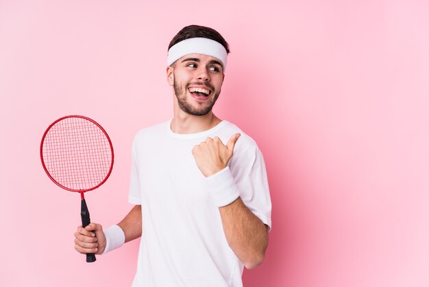 Jovem homem caucasiano jogando badminton isolado pontos com o dedo polegar afastado, rindo e despreocupado.