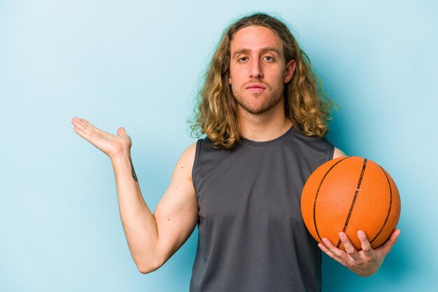 Jovem homem caucasiano jogando basquete isolado em fundo azul, mostrando um espaço de cópia na palma da mão e segurando a outra mão na cintura