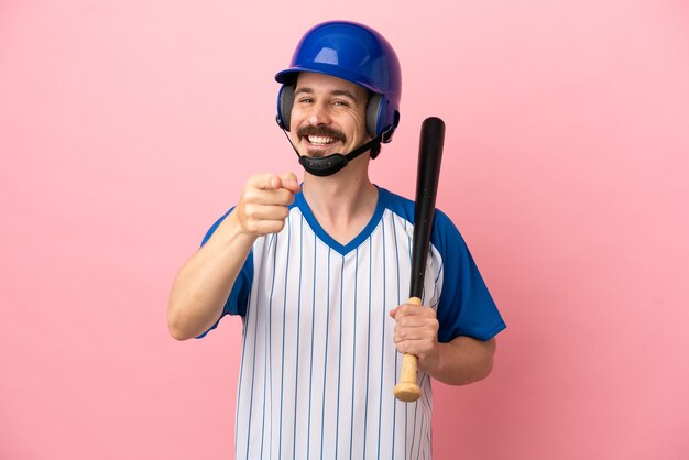 Jovem homem caucasiano jogando beisebol isolado em um fundo rosa apontando para a frente com uma expressão feliz