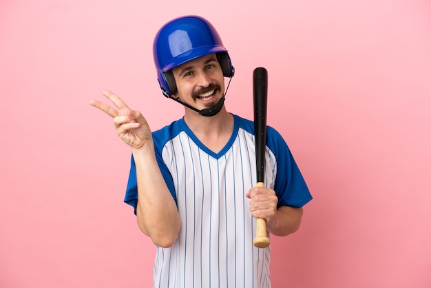 Jovem homem caucasiano jogando beisebol isolado em um fundo rosa sorrindo e mostrando sinal de vitória