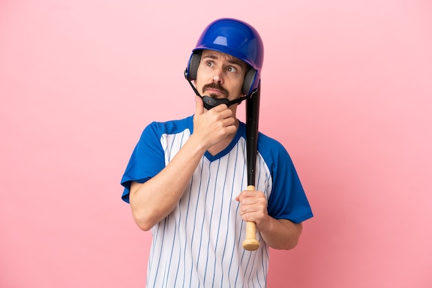 Jovem homem caucasiano jogando beisebol isolado em um fundo rosa, tendo dúvidas