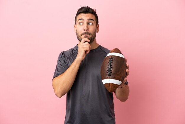 Jovem homem caucasiano jogando rugby isolado em um fundo rosa, tendo dúvidas e pensando