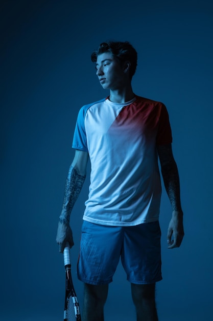 Jovem homem caucasiano jogando tênis isolado no fundo azul do estúdio em ação neon e conceito de movimento