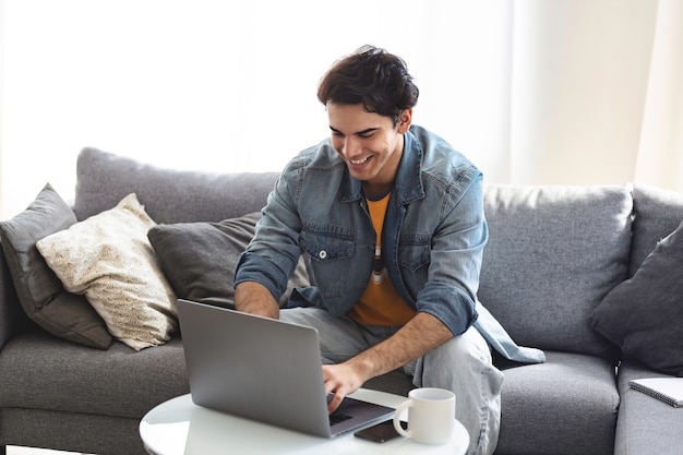 Jovem homem caucasiano olha para a tela do laptop falar em vídeo chamada online ou bate-papo nas redes sociais enquanto está sentado no sofá em casa. Conceito de tecnologia