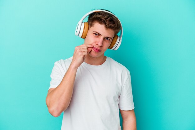 Jovem homem caucasiano ouvindo música com fones de ouvido isolados na parede azul com os dedos nos lábios, mantendo um segredo.