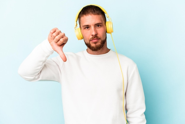 Jovem homem caucasiano ouvindo música isolada em um fundo azul, mostrando um gesto de antipatia, polegares para baixo. conceito de desacordo.