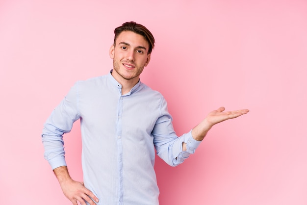 Jovem homem caucasiano posando em uma parede rosa isolada mostrando um espaço de cópia em uma palma e segurando a outra mão na cintura.