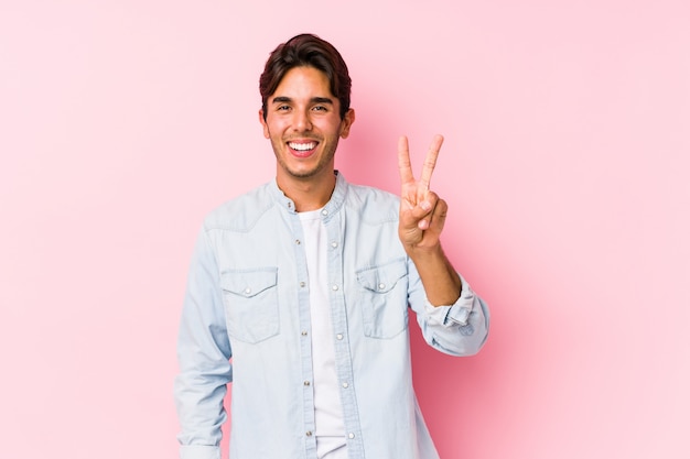 Jovem homem caucasiano posando em uma parede rosa mostrando sinal de vitória e sorrindo amplamente.