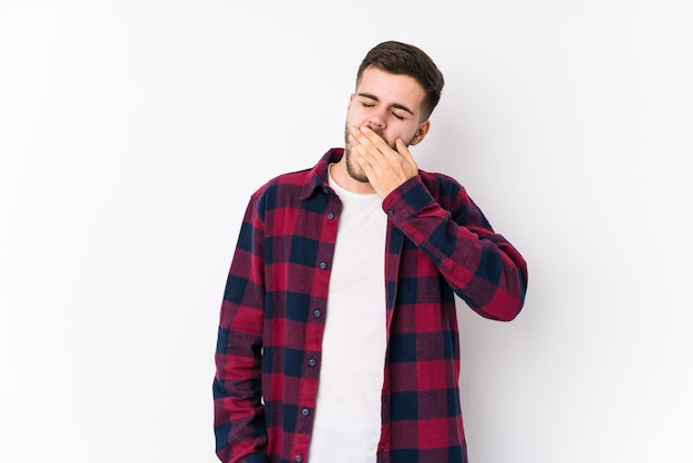 Jovem homem caucasiano posando em uma superfície branca isolada bocejando, mostrando um gesto cansado, cobrindo a boca com a mão.