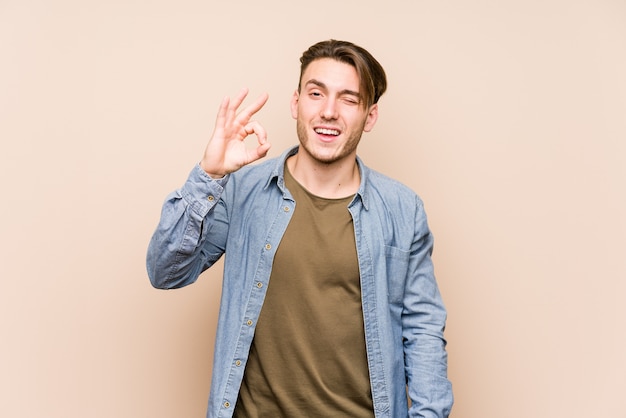 Jovem homem caucasiano posando isolado pisca um olho e mantém um gesto bem com a mão.