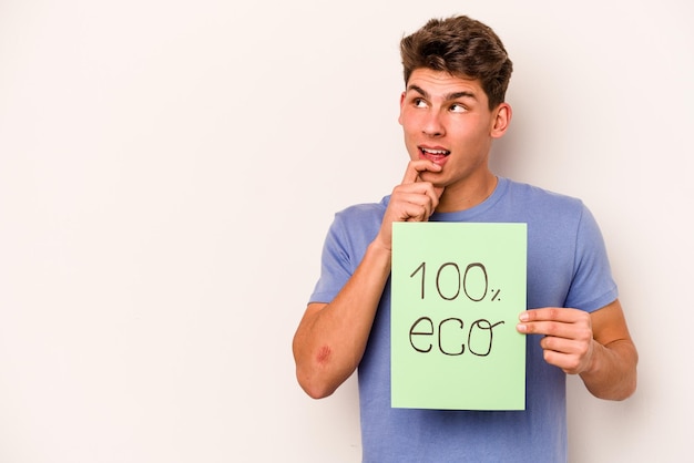 Jovem homem caucasiano segurando 100 eco cartaz isolado no fundo branco relaxado pensando em algo olhando para um espaço de cópia