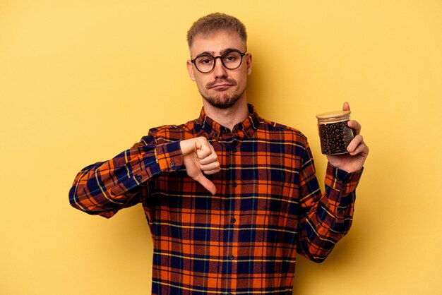 Jovem homem caucasiano segurando a jarra de café isolada em fundo amarelo, mostrando um gesto de desagrado, polegares para baixo. Conceito de desacordo.