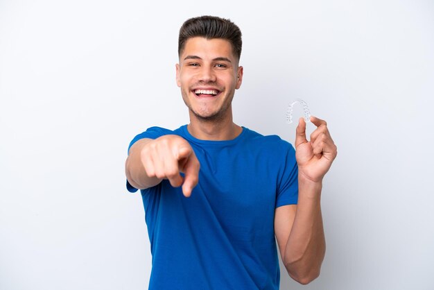 Jovem homem caucasiano segurando aparelho invisível isolado no fundo branco aponta o dedo para você com uma expressão confiante