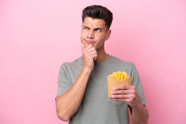 Jovem homem caucasiano segurando batatas fritas isoladas em fundo rosa com dúvidas e pensando