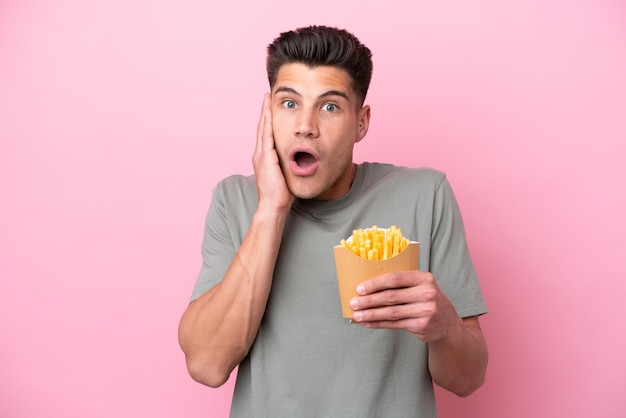 Jovem homem caucasiano segurando batatas fritas isoladas no fundo rosa com surpresa e expressão facial chocada