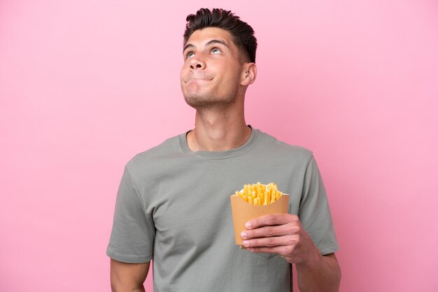 Jovem homem caucasiano segurando batatas fritas isoladas no fundo rosa e olhando para cima