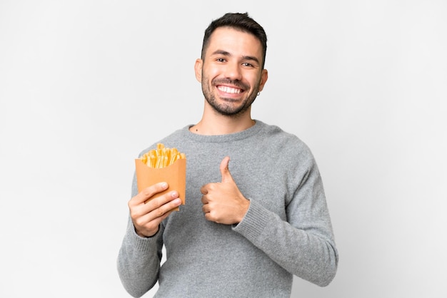 Jovem homem caucasiano segurando batatas fritas sobre fundo branco isolado, dando um polegar para cima gesto