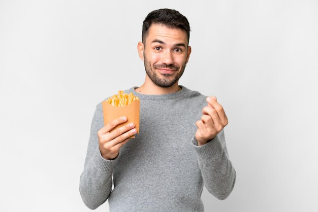 Jovem homem caucasiano segurando batatas fritas sobre fundo branco isolado fazendo gesto de dinheiro