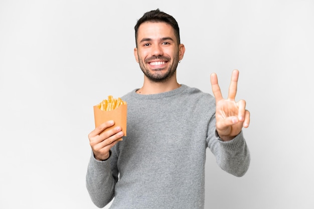 Jovem homem caucasiano segurando batatas fritas sobre fundo branco isolado, sorrindo e mostrando sinal de vitória