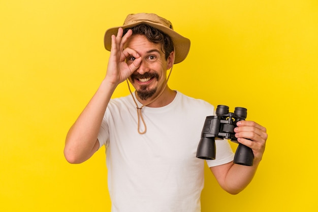 Jovem homem caucasiano segurando binóculos isolados em fundo amarelo animado, mantendo o gesto ok no olho.