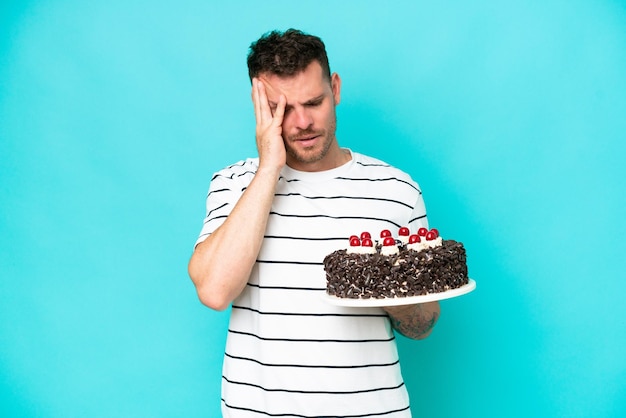 Jovem homem caucasiano segurando bolo de aniversário isolado em fundo azul com dor de cabeça