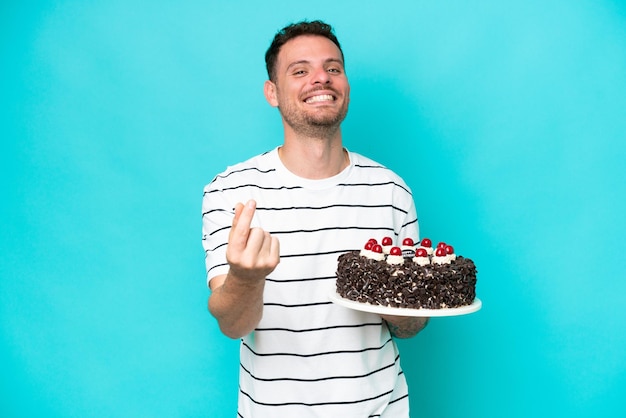 Jovem homem caucasiano segurando bolo de aniversário isolado em fundo azul fazendo gesto de dinheiro