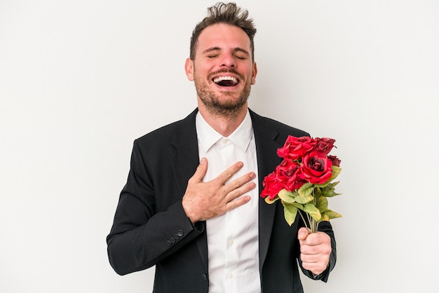 Jovem homem caucasiano segurando buquê de flores isoladas no fundo branco ri alto, mantendo a mão no peito.