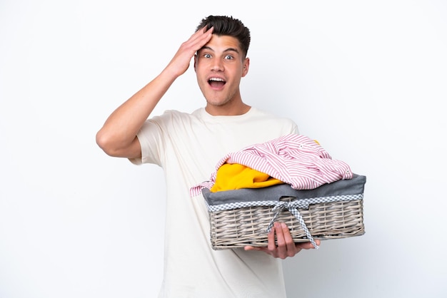 Jovem homem caucasiano segurando cesta de lavanderia isolada no fundo branco com expressão de surpresa