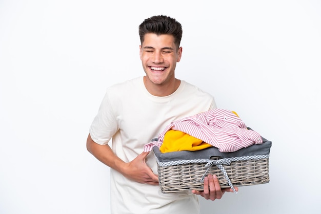 Jovem homem caucasiano segurando cesta de lavanderia isolada no fundo branco sorrindo muito