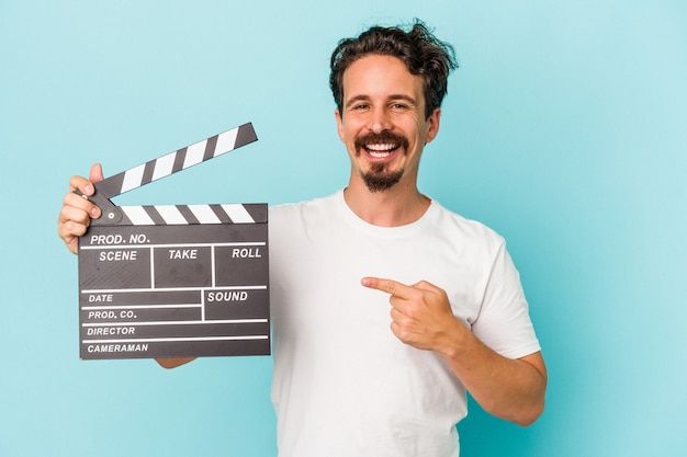 Jovem homem caucasiano segurando claquete isolada em fundo azul, sorrindo e apontando de lado, mostrando algo no espaço em branco.