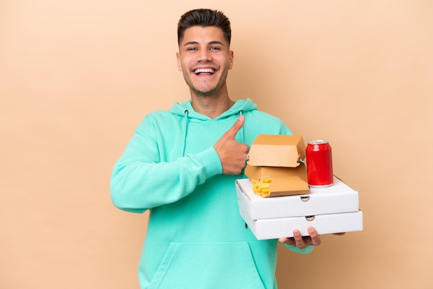 Jovem homem caucasiano segurando fast food isolado em fundo bege, dando um polegar para cima gesto