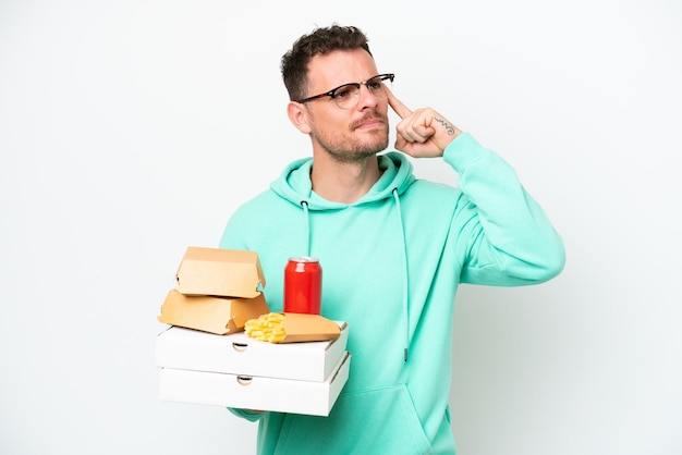 Jovem homem caucasiano segurando fast-food isolado no fundo branco com dúvidas e pensando