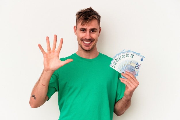 Jovem homem caucasiano segurando notas de banco isoladas no fundo branco, sorrindo alegre mostrando o número cinco com os dedos.