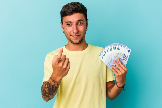 Jovem homem caucasiano segurando notas isoladas em um fundo azul, apontando com o dedo para você, como se fosse um convite para se aproximar.