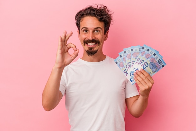 Jovem homem caucasiano segurando notas isoladas no fundo rosa, alegre e confiante, mostrando um gesto ok.