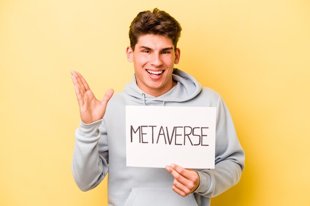 Jovem homem caucasiano segurando o cartaz do metaverso isolado em fundo amarelo, recebendo uma surpresa agradável animado e levantando as mãos