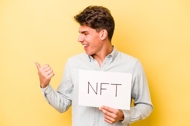 Jovem homem caucasiano segurando o cartaz NFT isolado em pontos de fundo amarelo com o polegar longe rindo e despreocupado