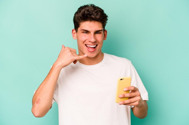 Jovem homem caucasiano segurando o celular isolado em fundo azul, mostrando um gesto de chamada de celular com os dedos