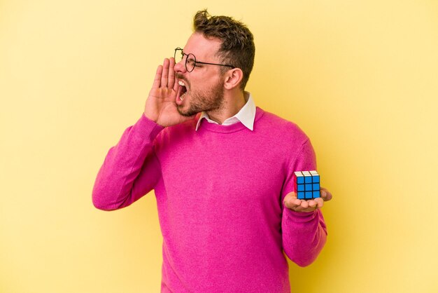 Jovem homem caucasiano segurando o cubo de Rubik isolado no fundo amarelo, gritando e segurando a palma da mão perto da boca aberta.