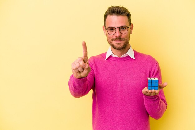 Jovem homem caucasiano segurando o cubo de Rubikâ € ™ s isolado no fundo amarelo, mostrando o número um com o dedo.