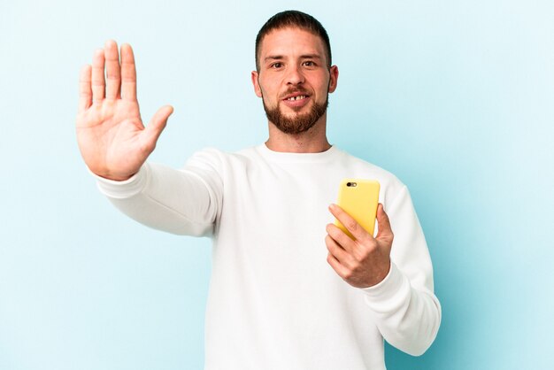 Jovem homem caucasiano segurando o telefone móvel isolado em um fundo azul em pé com a mão estendida, mostrando o sinal de stop, impedindo-o.