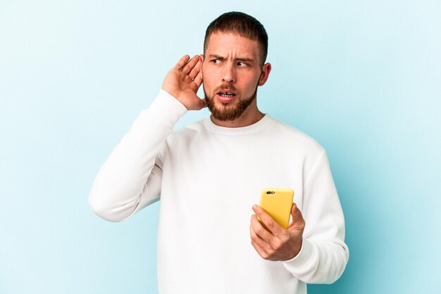 Jovem homem caucasiano segurando o telefone móvel isolado em um fundo azul, tentando ouvir uma fofoca.