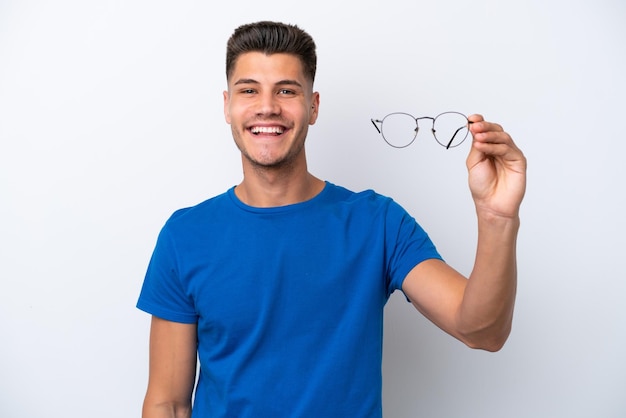 Jovem homem caucasiano segurando óculos isolados no fundo branco com expressão feliz